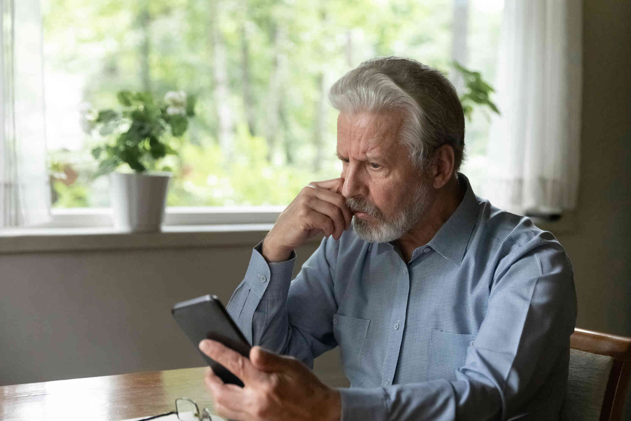 A middle aged man in a button down shirt sits at the table and looks at the cellphone in his hand with a worried expression.